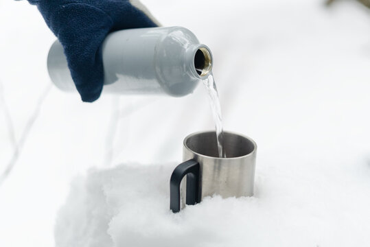 Closeup of a gloved hand pouring a drink from a bottle into a silver metal cup outdoors. Detail of winter beverage preparation with frosty background. Macro shot of liquid transfer in cold season