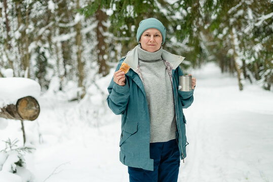 Adult woman enjoying a peaceful winter moment with a silver mug and pastry in snowy forest. Middle-aged lady in warm grey sweater standing in a calm relaxed pose outdoors. Tranquil quiet comfort