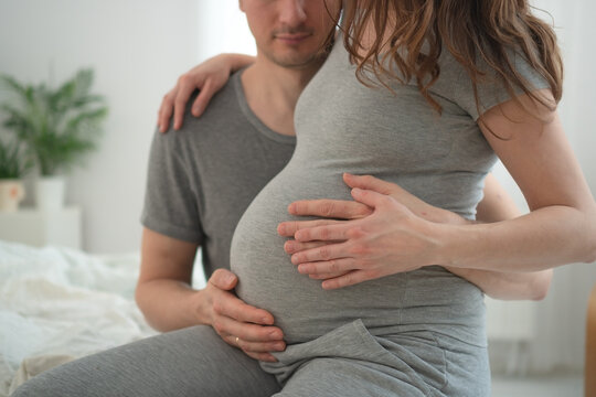 Pregnant woman and husband sitting on bed feeling baby move together in intimate moment. Expecting mother embracing partner while experiencing baby kick in cozy bedroom. Mom-to-be sharing romantic
