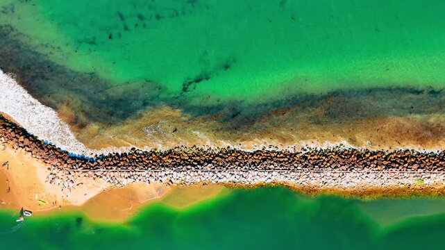 Mesmerizing sea-green waves hit the stone breakwater. People are having rest on the beautiful sandy beach. Top view.