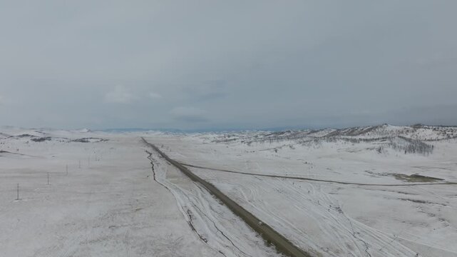 Drone footage of a long asphalt road cutting through snowy Tazheran Steppe in Irkutsk Oblast, Russia, near Lake Baikal, with hills and overcast sky