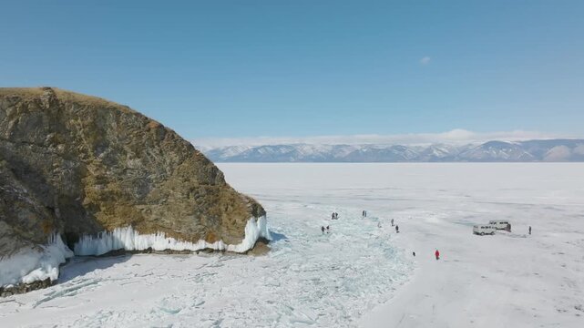 Aerial winter footage of Kharantsy Island rocky cape with icicles and hummocky ice on frozen Lake Baikal, Irkutsk Oblast, Russia, with tourists walking