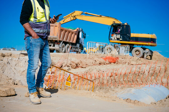 Construction worker using smartphone near excavator and truck