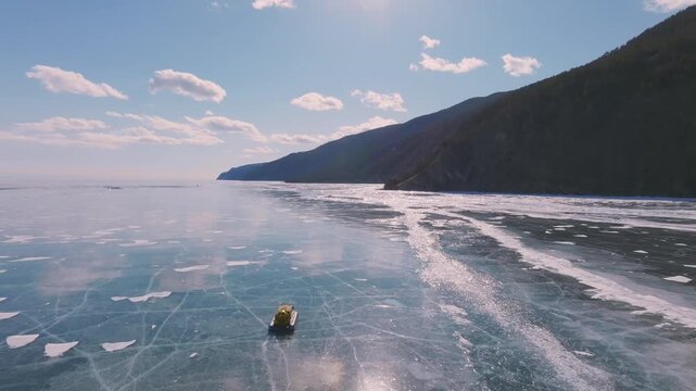 Aerial winter view of a hovercraft traveling across the clear ice of Lake Baikal near Listvyanka, Irkutsk Oblast, Russia, with mountains and blue sky