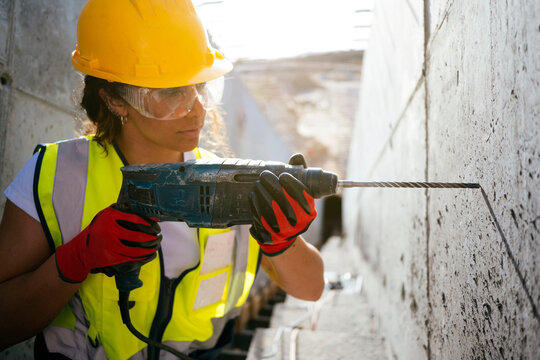 Female construction worker drilling concrete wall in safety gear
