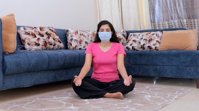 Pretty Indian girl meditating in Sukhasana (easy pose) for a healthy lifestyle. Sick young female doing breathing exercises while wearing a surgical mask during coronavirus pandemic in India