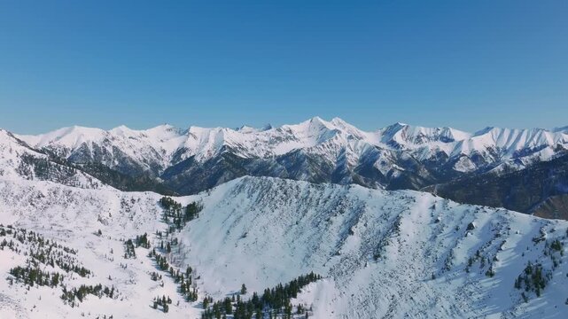 Aerial footage of snow-covered peaks and ridgelines around Peak Galina near Arshan, Republic of Buryatia, Russia, under clear blue sky, remote alpine wilderness