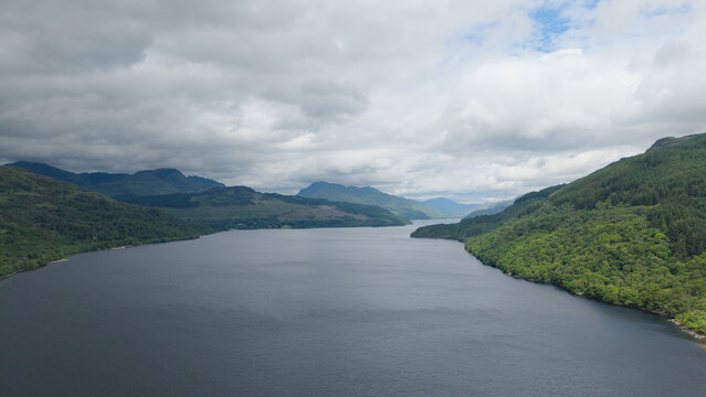 Breathtaking aerial panorama of Loch Lomond from Firkin Point, showcasing the vast lake surrounded by majestic mountains and lush green forests under a dramatic cloudy sky in Scotland.