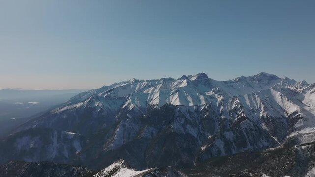 Wide aerial panorama of Galina Peak and rugged snow-covered ridges near Arshan village, Republic of Buryatia, Russia, under clear blue sky in winter daylight