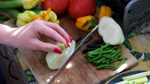 Fresh pattypan squash is being sliced on a wooden cutting board surrounded by cucumbers, green beans, pink tomatoes, flowers, and young zucchini. vegetables from the garden. 