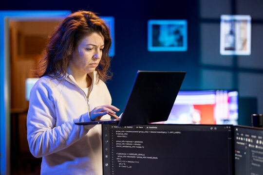 IT system administrator doing software debugging using laptop, checking glitches in lines of code displayed on desktop PC monitors. Woman standing in office at home fixing coding issues using notebook