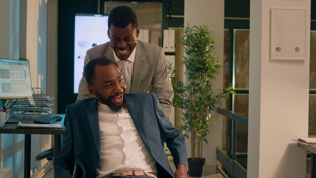 Jolly employee playing prank on unsuspecting colleague at work overnight, pushing his desk chair. Relaxed staff personnel having fun and laughing during nightshift in office