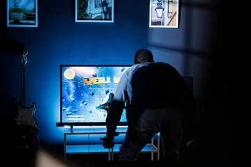 Guy wins video games online firing competition, overjoyed and pleased with his gaming activity. Young adult celebrates his victory in nighttime first person shooter championship. © DC Studio