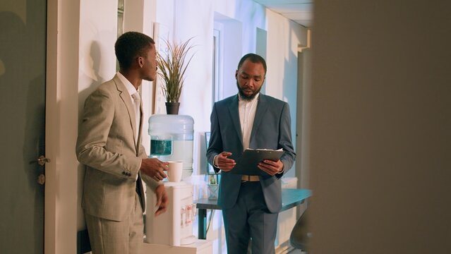 African american business manager checking up on employee project task progress during nightshift break. Relaxed office clerk enjoying cup of coffee, chatting with supervisor by the water tank