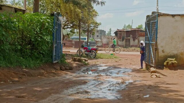 African girl in uniform standing near an open gate on a muddy village road, holding flowers and a backpack, with motorbikes and local houses in the background showing daily rural life.