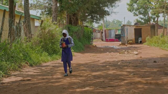 African girl in uniform standing near an open gate on a muddy village road, holding flowers and a backpack, with motorbikes and local houses in the background showing daily rural life.