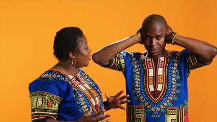 Obraz premium Furious girlfriend shouting at her boyfriend and fighting, having disagreement over orange background. Frustrated man covering his ears and trying to ignore his wife yelling in studio.