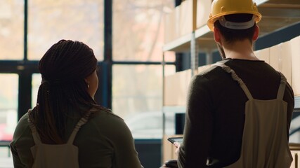 Proficient warehouse employees preparing orders for pickup, walking through storage building aisles and using digital device to check shipping info while scanning labels on cached items © DC Studio