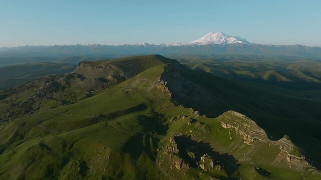 Drone footage of green cliffs and rolling hills of Bermamyt Plateau with snowcapped Mount Elbrus in the distance, Karachay-Cherkessia, Russia