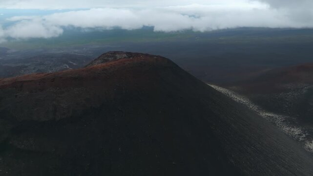Aerial footage of dark volcanic cinder cone and lava flows in Tolbachinsky Dol, Kamchatka Krai, Russia, with dramatic low clouds and vast barren landscape