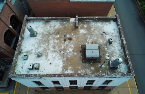 Aerial view of old commercial building roof. White flat roof shows signs of wear and tear, with pipes and vents visible. Adjacent brick structure suggests urban or industrial setting.