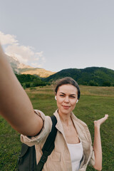 Woman takes a casual outdoor selfie in a green countryside field with a backpack, smiling under a...