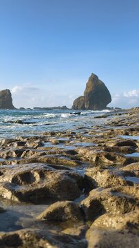 Slow Motion of Papuma Beach rocky textures with clear tide pools, featuring ocean waves crashing against a massive sea rock under a blue sky. Jember, Indonesia