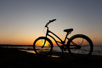 Naklejka premium Minimalist shot of a vintage cruiser bicycle silhouette standing on a sandy shore against a dramatic orange sunset sky and calm sea waves.