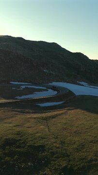 Vertical Screen: Car navigates curved section of Beartooth Highway in Montana USA during golden hour. Snow patches dot the rocky alpine terrain as vehicle rounds hairpin turn with guardrails.