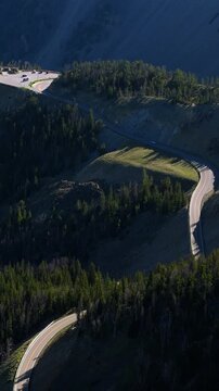 Vertical Screen: Beartooth Highway winds through rugged Montana USA mountain terrain with dramatic switchbacks cutting across forested slopes dotted with evergreen trees and parking area visible.