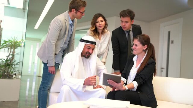 A Middle Eastern man in traditional attire and white-collar professionals sit on a couch in a sleek office, reviewing documents together.