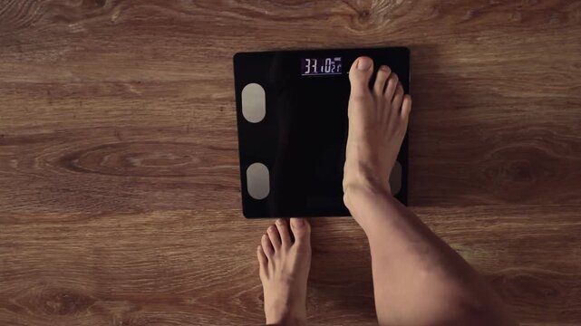 POV - top point of view video from above: woman bare feet stepping on black digital floor scales, she weighing, checking herself at home. Measuring weight, control, wellness, diet body care concept