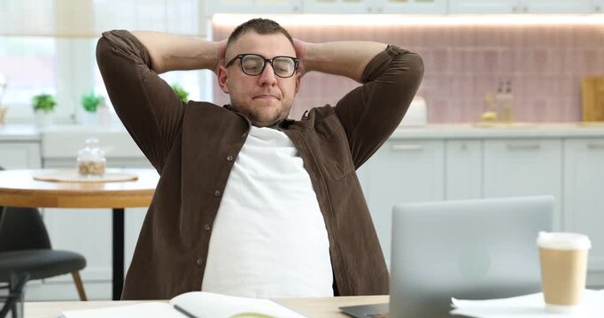 Workaholic. Overwhelmed man working with laptop and stretching at wooden desk in kitchen, camera moving
