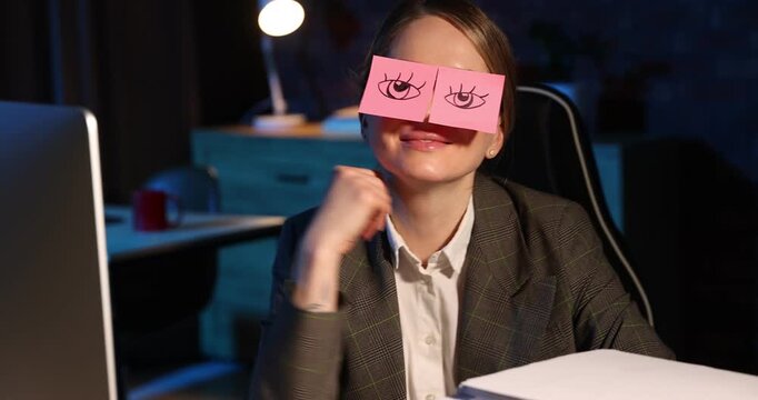 Workaholic. Smiling woman with fake eyes drawn on sticky notes between stack of documents and computer at desk indoors at night. Camera moving forward