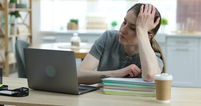 Workaholic. Overwhelmed woman at wooden desk with takeaway cup of coffee, laptop and stationery in kitchen. Camera moving forward