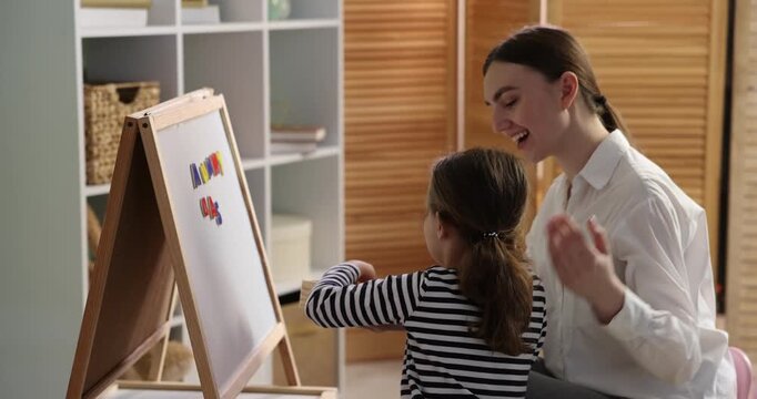 Speech therapist teaching girl alphabet with magnetic letters near whiteboard indoors