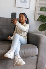 Obraz premium Asian female child sitting on gray suede couch at home holding black smartphone, wearing cream top