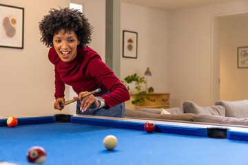 Fototapeta premium African American woman wearing red top leaning over blue-felt pool table at home, holding cue