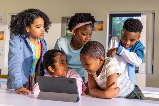 Diverse children gathering around tablet in folio on white classroom table, leaning forward