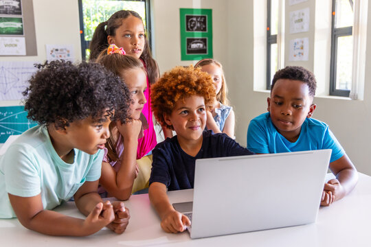 Diverse school-aged children leaning around silver laptop at table in classroom with math posters