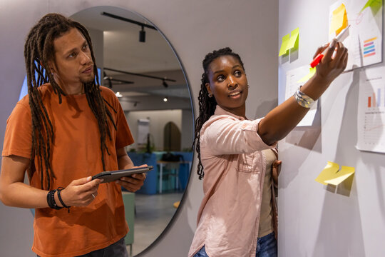 Diverse coworkers marking office wall charts woman in pink adding notes man with tablet, copy space
