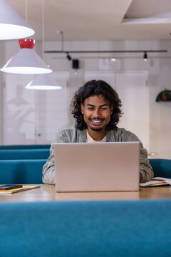 Indian man in his twenties wearing light jacket sitting at table in co-working space using laptop