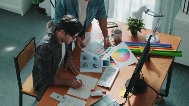 Top view of manager writing idea while team standing and giving comment. Professional designer design logo while looking at color palette and writing idea down in paper with color marker. Symposium.
