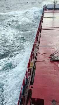 Vertical shot of large ocean waves crashing against the side of a cargo ship during a storm at sea.