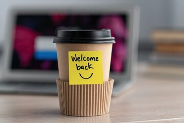 Fototapeta premium Coffee cup with welcome back sticky note in office, laptop background, smiley face