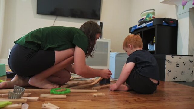 Mother and child work together on the floor, assembling wooden train tracks while colorful toys are scattered around in a bright living room with a television and storage units visible in background