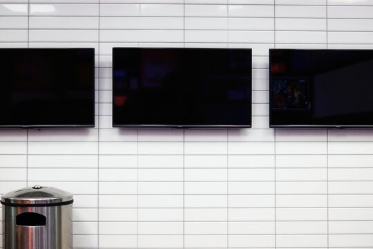 Three large flat screen TVs mounted on a white tiled wall, one displays menu content. A metal rubbish bin sits below. Modern restaurant digital information display.