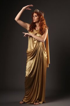 Woman in golden toga poses with crown. She has red hair, graceful hand gestures. Studio portrait. Ancient Greek goddess style with elegant draping.