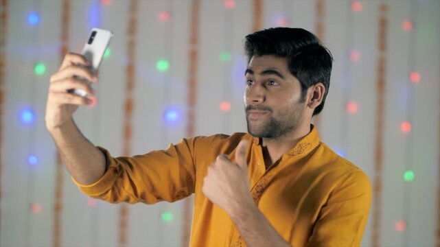 A handsome Indian man in a kurta takes selfies from different angles . Young Asian man traditionally dressed trying different selfie poses in a house decorated with lights and flowers