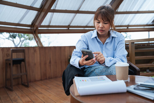 Woman in blue shirt is focused on her phone while sitting at wooden table with financial documents and coffee cup, engaged in data analysis and planning discussion